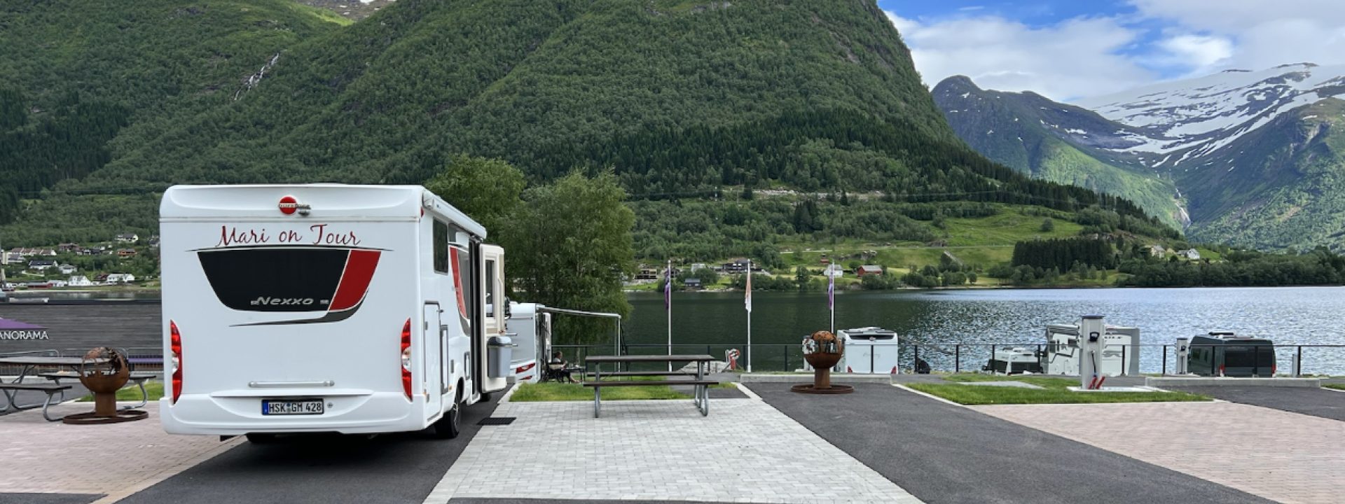 Bidet viser en hvit bobil parkert på en asfaltert parkeringsplass med en spektakulær utsikt over et vann og fjellandskap.